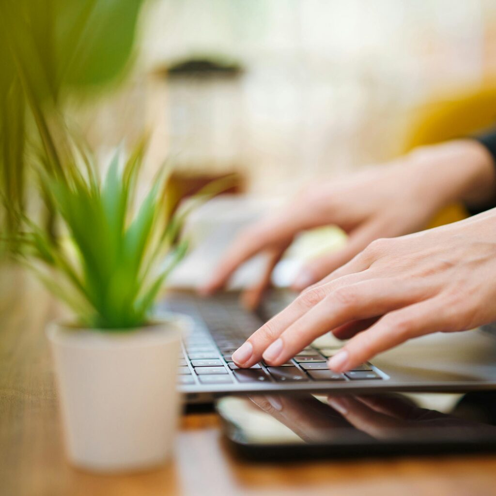 Detailing a moment in the daily operations of a social media marketing agency, this image showcases a close-up view of a professional's hands actively typing on a laptop, with a small potted plant adding a natural element to a focused and modern remote work setting.